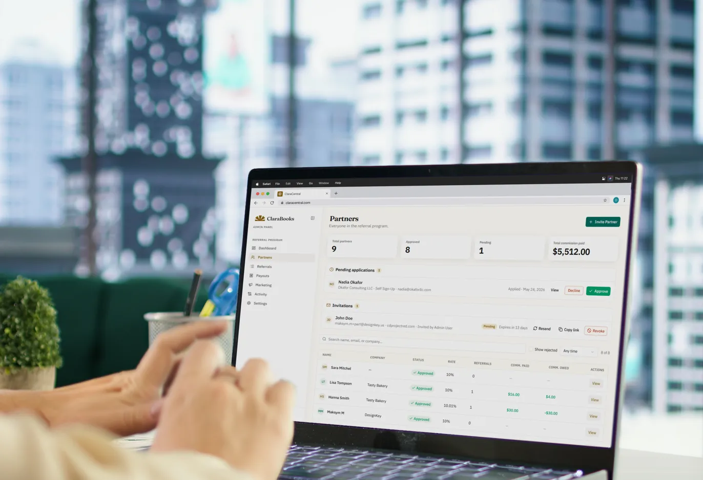 A laptop on a desk beside a city window, showing the admin Partners panel: totals for partners, approvals and commission paid, a pending application awaiting a decision, an outstanding invitation, and the partner list with each partner’s rate and commission owed.