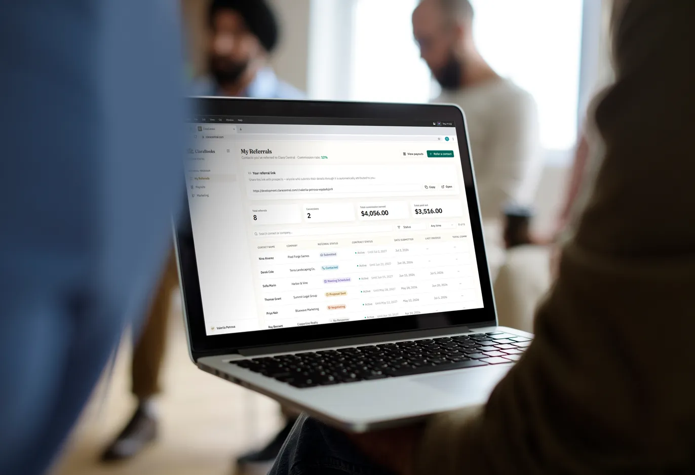 A laptop held on someone’s lap in an open-plan office, showing the partner portal’s My Referrals page: the partner’s own referral link, totals for referrals, conversions and commission earned, and a table of the contacts they have referred with the status of each.